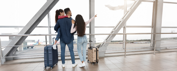 A family of three looks out of an airport window at planes departing in the background.